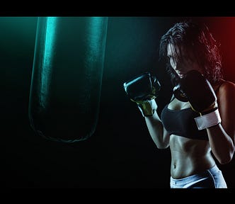 A woman in shadow wearing a black sports bra, shorts, and boxing gloves, holding her hands up ready to fight.