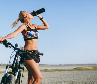 Young woman in sports outfit drinking water from bottle while holding her bike. She appears to be in a barren landscape. She is wearing shorts and colourful sports top. Her blond hair is tightly knotted in a pony tail. Her sunglasses rest on her head as she drinks.