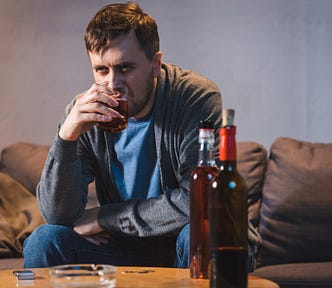 man with beard on couch drinking alcohol from glass two bottles of liquor on table