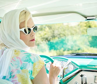 A blonde woman with sunglasses drives a 1950s era car