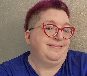 An obese white woman with red cat-eye glasses and a short purple mullet smiles at the camera.