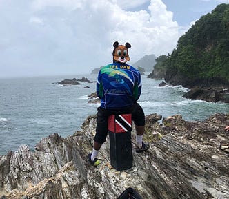 Leevan in a blue Trail Trekkers TT shirt with his name on it. His back faces us and he sits on a stool with the Trinidad and Tobago flag at the top of the stool. The stool is near the edge of a precipice and he’s facing the sea on a cloudy day.