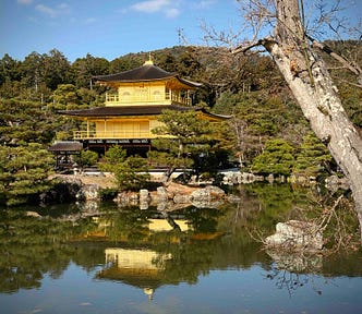 Kinkaku-ji (Golden Pavilion) in Kyoto, Japan, a three-story Zen Buddhist temple with gold-leaf-covered upper floors, reflected in a tranquil pond and surrounded by lush greenery. A large tree trunk appears in the foreground, adding depth to the serene landscape.