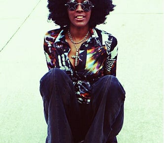 Woman with afro hair and star patterned sunglasses wears multicolour shirt with large collar, cross necklace and dark flared trousers — 1970s fashion