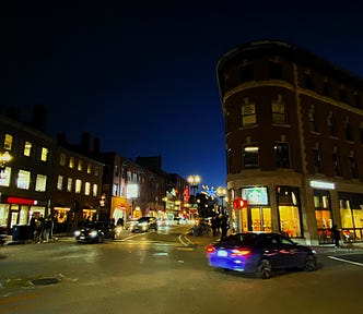 Street scene at night. Non-descript buildings with their lights on and cars passing on the street, a few pedestrians are walking on sidewalks.