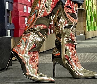 The author is wearing 5 inch tall golden stilettos. Bulky men’s jeans are being lifted up to reveal them in a shoe store with lots of stacked red and green shoe boxes in the background.