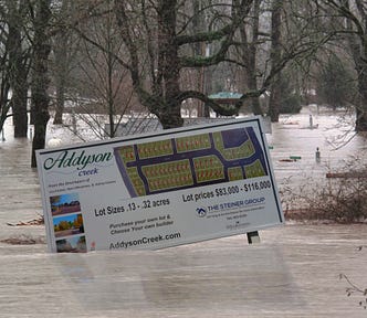 A leafy suburb, flooded to the roofline. In the foreground is a sign advertising a new subdivision, askew and partially submerged. Image: Bdelisle (modified) https://en.wikipedia.org/wiki/File:Snoqualmie_area_flood.jpg CC BY 3.0 https://creativecommons.org/licenses/by/3.0/ Rick Obst (modified) https://www.flickr.com/photos/discoveroregon/28381003281/ CC BY 2.0 https://creativecommons.org/licenses/by/2.0/