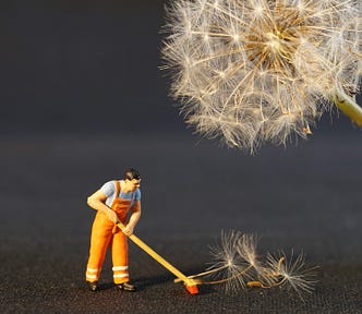 Toy working in high-visibility clothing sweeping up the seeds of a dandelion. The dandelion is looming over the worker.