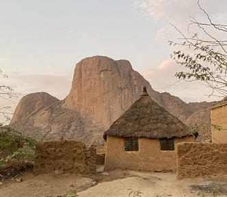 A mud and grass thatched hut sits on a hill in front of unique mountains.