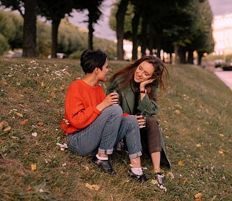Two friends sitting and talking on a grassy hill, drinking coffee from paper cups. One wears a vibrant orange knit sweater and jeans, while the other wears a long green coat, capturing an intimate, real-world moment of human connection.