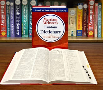 Two dictionaries, one standing that reads “Merriam-Webster’s Fandom Dictionary”, one lying open and flat in front of it.