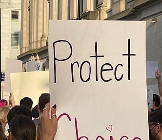 A hand with two rings on it is holding a sign that says “Protect Choice” in the foreground of a crowd in front of an official looking stone building.