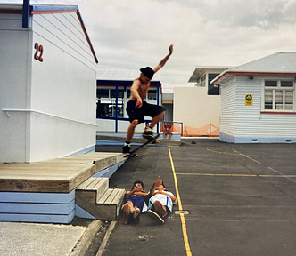 A skateboarder is captured in mid-air, he’s halfway through an olly – jumping off  two stairs and over two humans (his friends). It’s a make or break moment. He’s mid-flight directly above his two friends, who are lying down beneath him. One is throwing a shaka, the other is giving the thumbs up. Two younger kids watch the action from the distant background. They have no shoes. They wait to see if he lands it, or if he crashes into his mates.