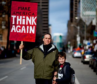 A man holding a sign saying “Make America Think Again,” and standing next to a small boy.