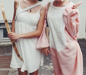 Two women standing side-by-side, one in a white dress and carrying a wooden bow. The other in a Pink Panther onesie.