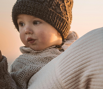 Toddler is bundled in crocheted hat and knit sweater and looking over mom’s shoulder who is carrying him and walking away from the camera. They are both bundled for the snow.