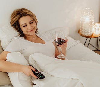 A smiling woman lying in bed, holding a glass of wine and a TV remote, with candles and a coffee mug to her left.