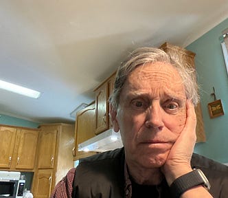 A man with graying hair and a concerned expression rests his head on his hand while sitting in a kitchen with wooden cabinets. He wears a black vest over a checkered shirt and a smartwatch on his wrist. The background includes a stove hood, a refrigerator with magnets, and a window with a rolled-up shade. The lighting is soft and natural.