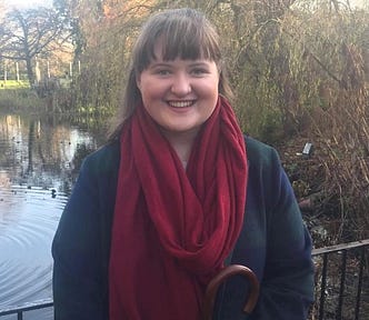 The author, smiling, wearing a red scarf, against the backdrop of a pond or stream and a thicket of trees.