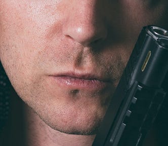 The lower half of a man’s face with a pistol resting on his cheek.