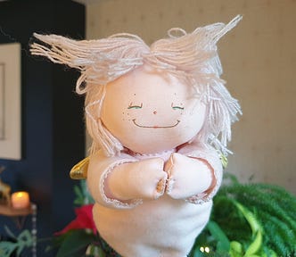 Photo of a pink Christmas tree fairy. She has the most beautiful smile and her hands are clasped together in front of her. Her hair is made of pink cotton threads and it is sticking out at funny angles. The blurred background shows green foliage, a poinsettia, a candle and other Christmas decorations.