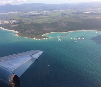 A plane wing out a plane window flying over the turquoise sea and white beaches of an island.