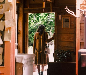 a woman walking out the door of a house, she is wearing a long brown coat and looking down