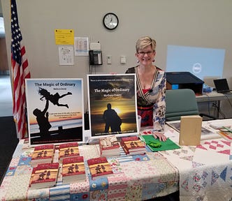 The author, Melissa Gouty, is smiling and standing behind a quilt-covered table that has two posters of her book, The Magic of Ordinary, and copies of her book.