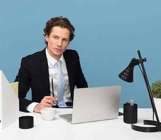 Dark-haired man dressed in black suit with white shirt and blue tie, sitting at a modernist desk with laptop, black lamp, and plant, against a bright blue background.