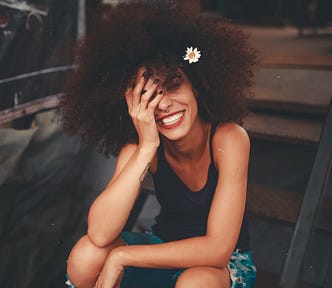 A pretty girl with a flower adorning her curly hair sits with a big smile on her face. She looks like she’s having an epiphany, reflecting at what she’s been through in life and she’s happy in the moment.