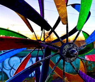 A closeup of a windmill with many colors in front of a sunset and a fence covered in graffiti writing something indeciferable.