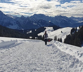 A woman walks on the snow, with the Swiss Alps on the horizon. The landscape is full of snow. The sky is blue, dotted with scattered white clouds.