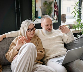 An older couple sitting on the couch waving “hello” at a laptop computer screen.