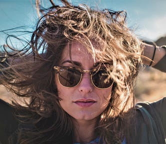 girl with brown hair wearing sunglasses and dark top with hands behind her head
