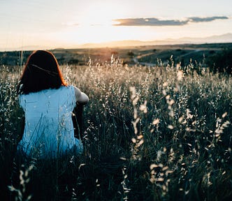 Woman watching sunset, sits in a field, her back faces the camera.