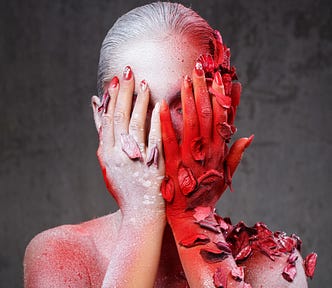 Headshot of a woman with her hands covering her face. She is spray-painted white on one side and red on the other. There are also leaves clinging to her. They too are spray painted.