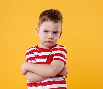 A grumpy-looking young boy in a red and white T-shirt against a yellow background. He has his arms folded across his upper body and looks quite cross.