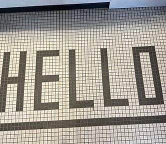 The image shows a tiled floor with the word “HELLO” spelled out in large, black, capital letters. The tiles are small and square, with a white background and black lines forming the letters and a border around the word. The word “HELLO” is centered within the border.