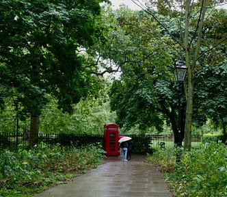 Three tourists under umbrellas look at a red telephone booth outside a park