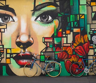 Wall mural showing multicolored shapes around a woman’s face with two bicycles leaning on the wall.