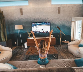 A woman sitting on a sofa watching a ice hockey game, raising her hands up in the air.