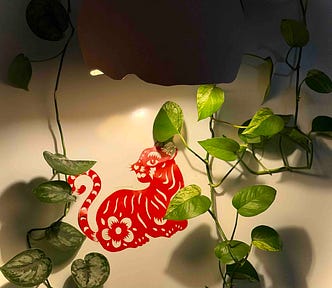 A red paper‑cut cat with floral patterns hangs on a light wall, surrounded by trailing green pothos vines that frame the artwork with soft, natural curves.