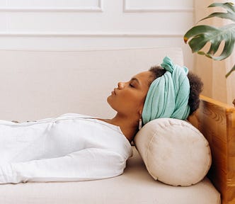 A woman in a white top with a light turquoise reclines on a white couch with her eyes closed.