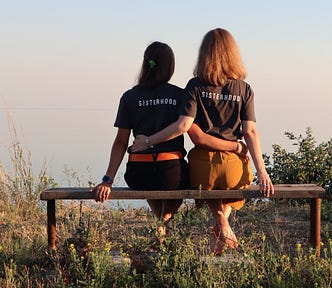 A rear view of two woman sitting on a bench. They each have an arm around the other’s waist. They are both wearing black t-shirts with the word ‘sisterhood’ on the back.