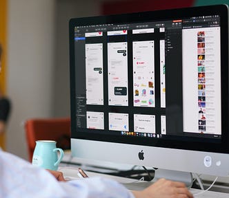 computador da apple, é um computador desktop de tela grande. Na tela aparece alguns elementos de design e componentes de design. Na imagem aparece recortado uma mão de homem de pele caucasiana. Ao redor tem uma jarra azul.