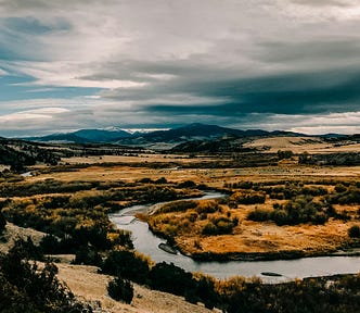 A panorama of Montana ranchlands showing a river winding throw pasture lands with snow-capped mountains in the background.