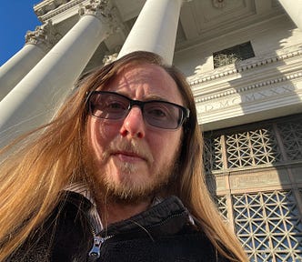 The author appears to be looking down at the audience. He has long brown hair, sunglasses, a beard, and a mustache tinged with white showing his age. Wrinkles are furrowing his forehead showing that he’s serious. Behind him are the pillars of a courthouse. An iron door is behind him. Above it are Greek letters. The sky is a striking blue in the background.