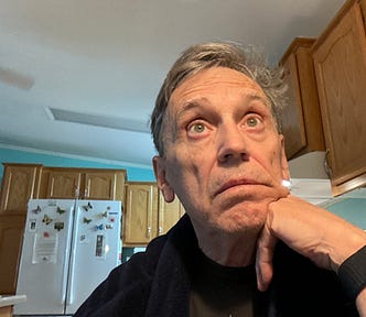 A middle-aged man with graying hair is looking upward in a kitchen, perhaps thinking of something. The background features wooden kitchen cabinets, a refrigerator with colorful butterfly magnets, a microwave, and a light blue wall.