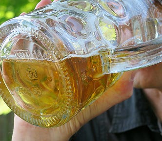 Most of the image is taken up by a large half-filled beer glass, tilted to someone’s mouth. You can only see part of their face on the right of the image, and part of their hand holding the glass.