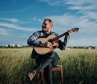 A happy man in a denim jacket playing his guitar in lush fields